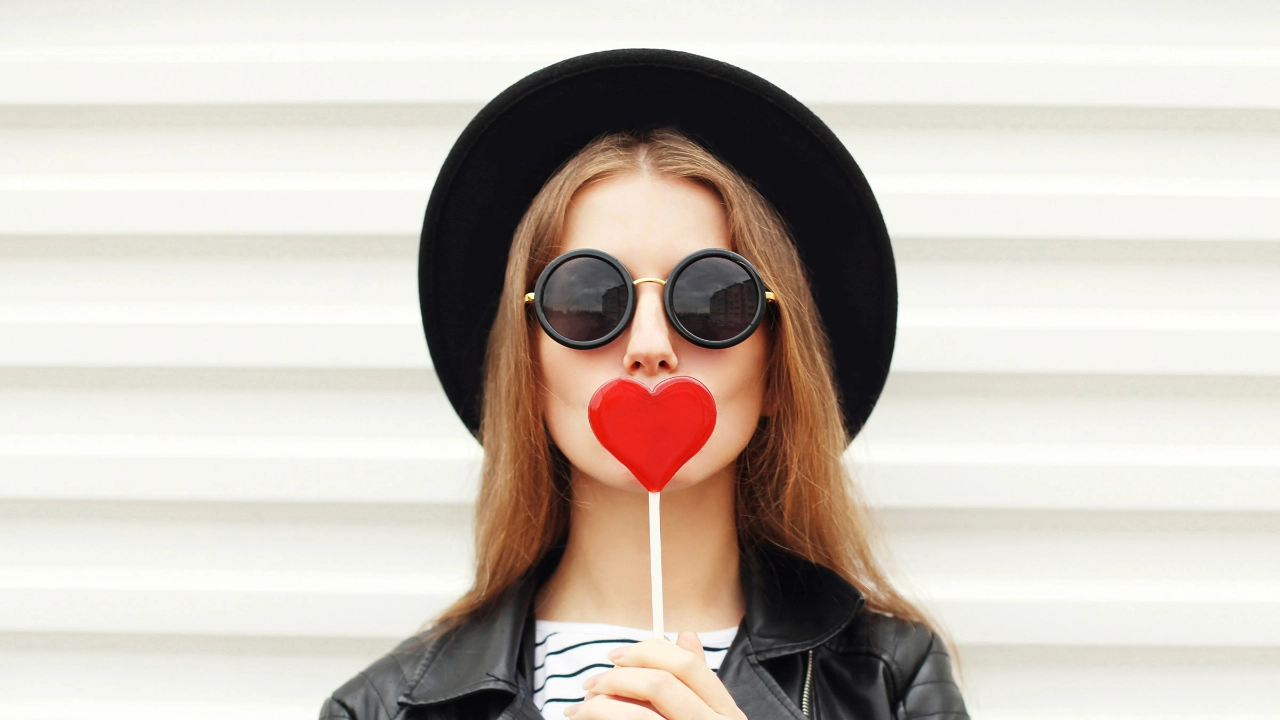 Junge Frau mit Sonnenbrille hält sich einen Lolly in Herzform vor den Mund.